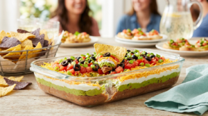 A clear glass dish of 7 layer dip featuring distinct layers of refried beans, guacamole, sour cream, and shredded cheese, finished with fresh diced tomatoes, black olives, and green onions, with a tortilla chip scooping out a bite.