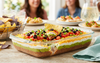 A clear glass dish of 7 layer dip featuring distinct layers of refried beans, guacamole, sour cream, and shredded cheese, finished with fresh diced tomatoes, black olives, and green onions, with a tortilla chip scooping out a bite.